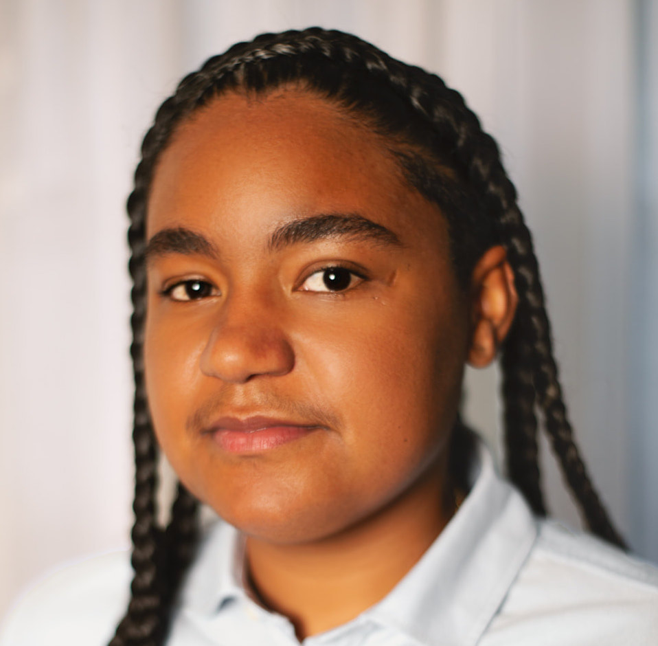A photo of Ana Conner, a Black gender non-conforming person with black braids going over their shoulders. They're wearing a light collared shirt.